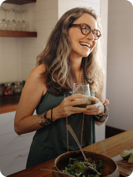 Mulher de óculos e cabelos grisalhos sorrindo enquanto segura um copo com suco proteíco durante o almoço saudável.